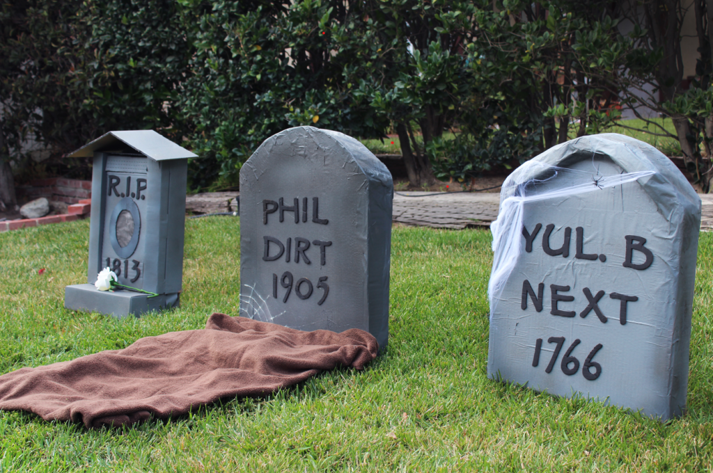 Three humorous cardboard tombstones on a grass lawn, decorated with cobwebs, a brown cloth, and a white flower.